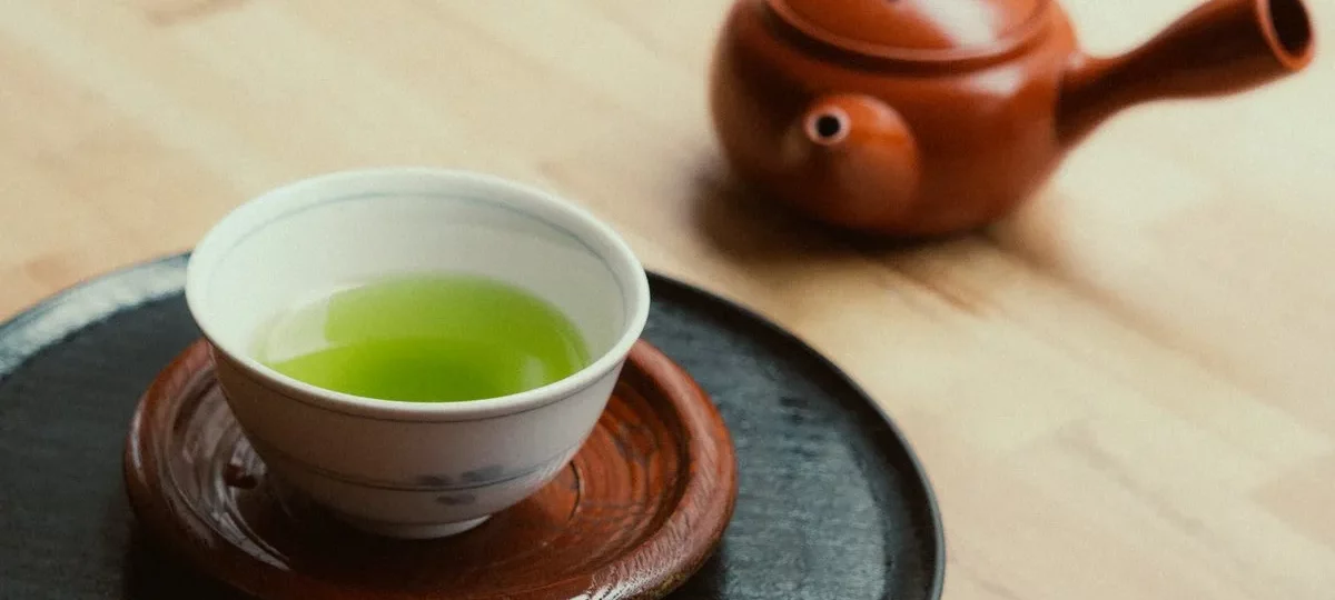 Bol de thé vert à la robe vert vif et théière japonaise en terre rouge sur une table en bois clair