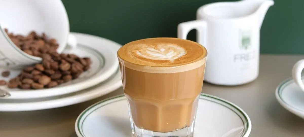 Café noisette dans un petit verre sur soucoupe, pot à lait et grains de café sur une table de café
