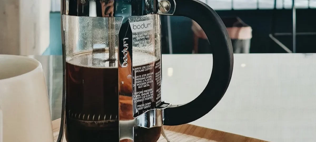 Cafetière à piston Bodum en verre remplie de café, posée sur un plateau en bois près d'une baie vitrée