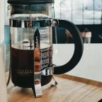 Cafetière à piston Bodum en verre remplie de café, posée sur un plateau en bois près d'une baie vitrée