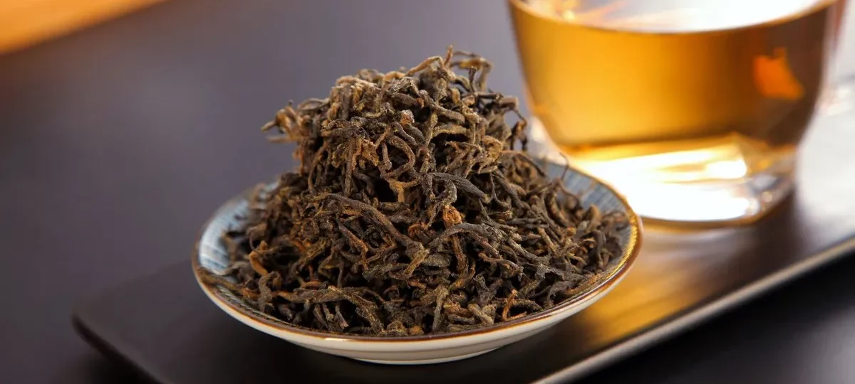 Feuilles de thé noir torsadées dans une coupelle devant une tasse de thé infusé