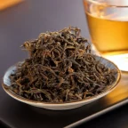 Feuilles de thé noir torsadées dans une coupelle devant une tasse de thé infusé