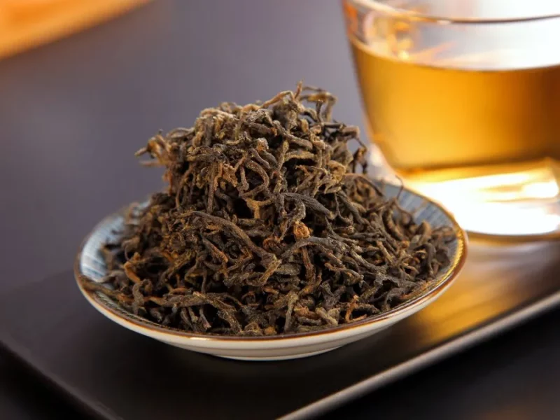 Feuilles de thé noir torsadées dans une coupelle devant une tasse de thé infusé