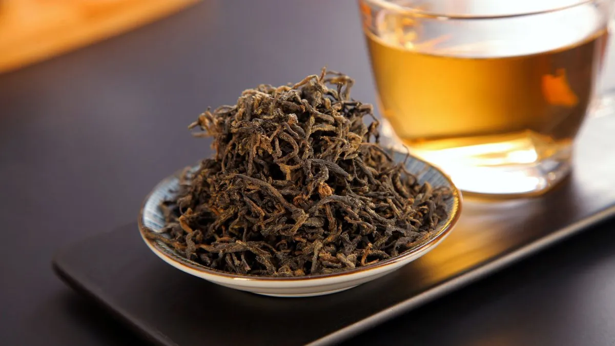 Feuilles de thé noir torsadées dans une coupelle devant une tasse de thé infusé