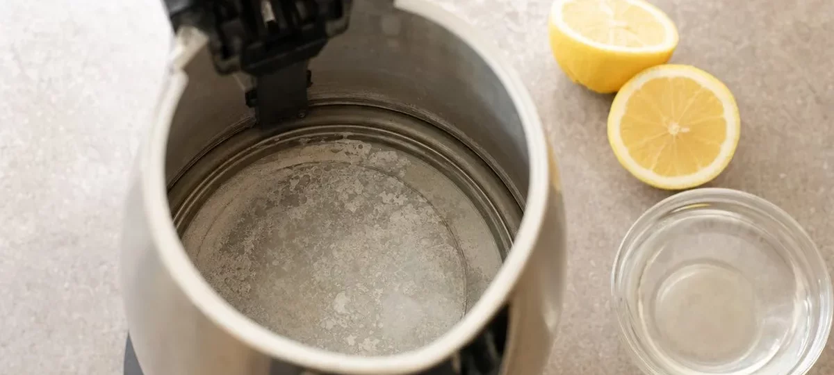 Intérieur d'une bouilloire inox couvert de calcaire, à côté d'un citron coupé et d'un bol de vinaigre blanc