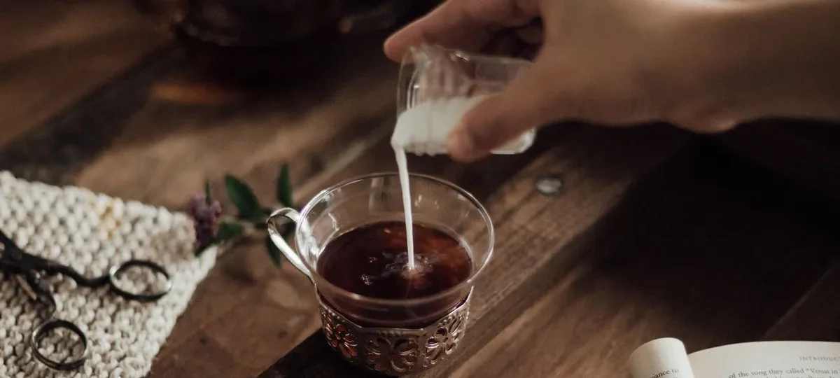 Lait versé dans une tasse en verre de thé noir, théière en verre en arrière-plan sur table en bois