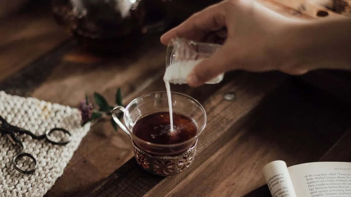 Lait versé dans une tasse en verre de thé noir, théière en verre en arrière-plan sur table en bois