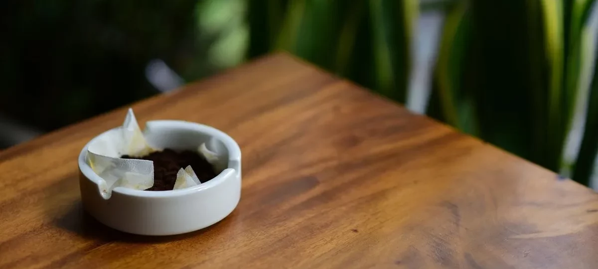 Marc de café usagé dans une petite coupelle blanche sur une table en bois, plantes vertes derrière