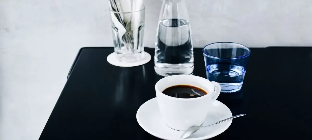 Tasse de café americano noir sur soucoupe à côté d'une carafe d'eau, sur une table noire