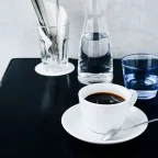 Tasse de café americano noir sur soucoupe à côté d'une carafe d'eau, sur une table noire