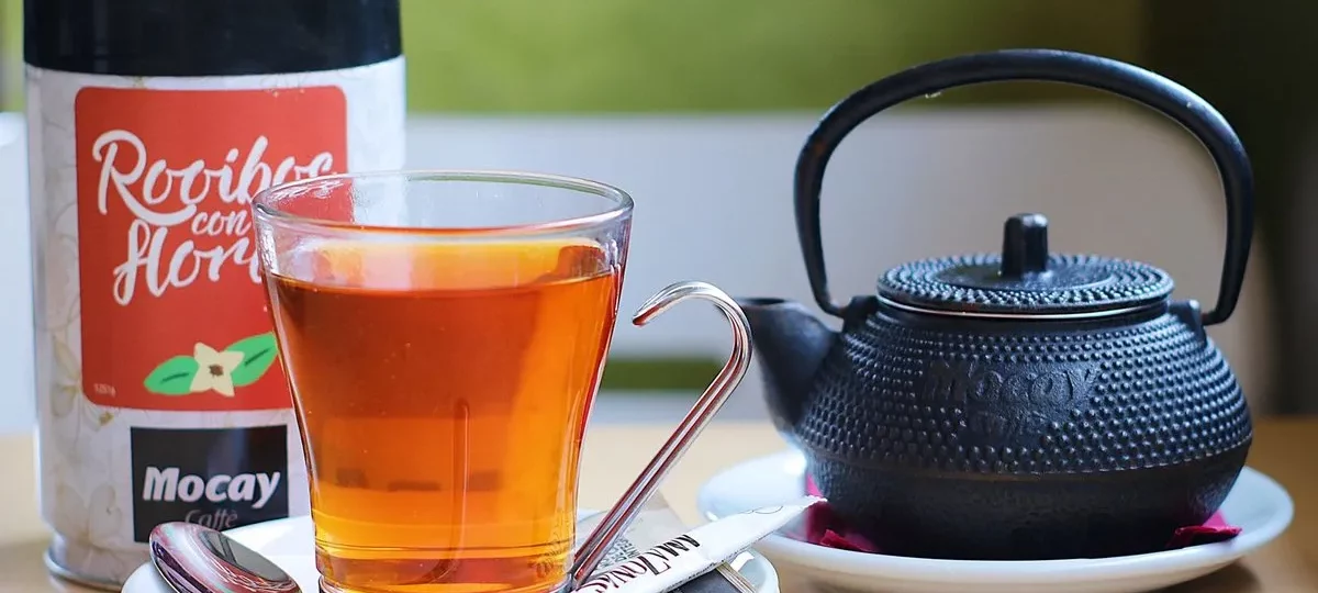 Verre de rooibos infusé à la robe rouge-orangé, à côté d'une boîte de rooibos et d'une théière en fonte