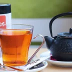 Verre de rooibos infusé à la robe rouge-orangé, à côté d'une boîte de rooibos et d'une théière en fonte
