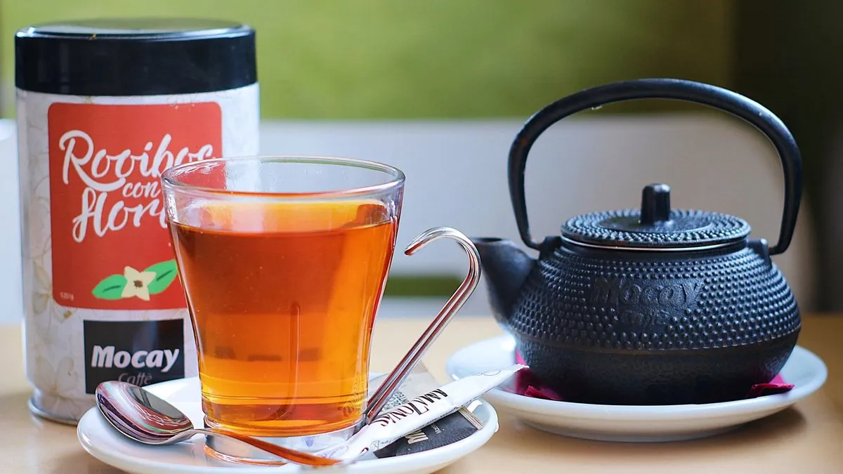 Verre de rooibos infusé à la robe rouge-orangé, à côté d'une boîte de rooibos et d'une théière en fonte